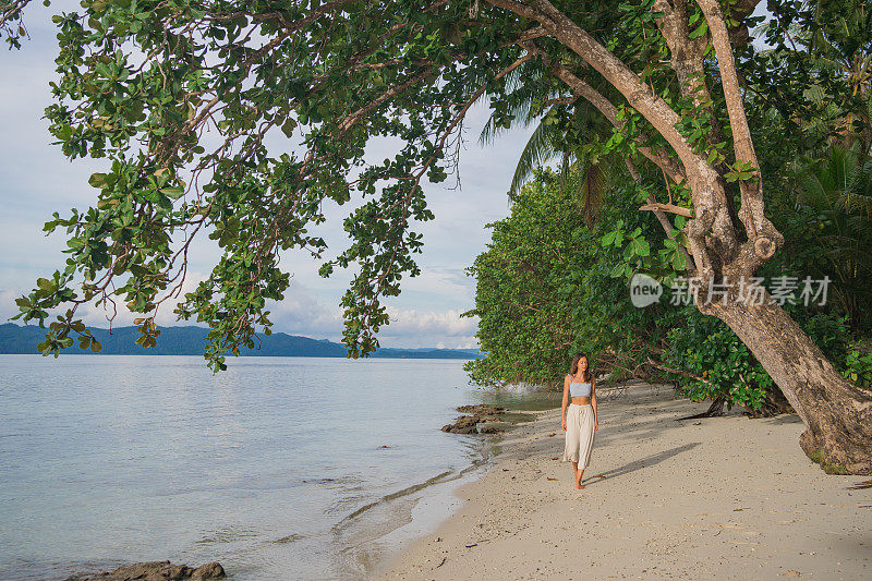 年轻女子走在树下的白色沙滩上
