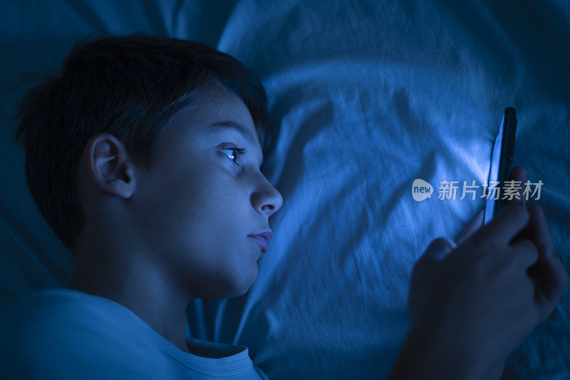男孩晚上在床上玩智能手机。10岁男孩睡觉前玩手机。孩子在自己的房间里不受控制地使用智能手机。本空间