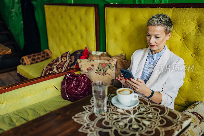 一位留着短发的女商人坐在咖啡店里喝咖啡休息时正在玩手机