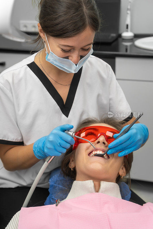 女牙医在口腔检查期间为病人洗牙。一名年轻女子戴着安全眼镜坐在牙科椅上看牙医。垂直图