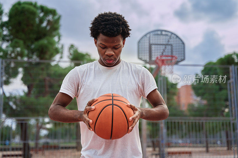 年轻英俊的非裔美国男子篮球运动员在户外双手持球。有篮球场的城市背景。男篮球运动员的肖像。