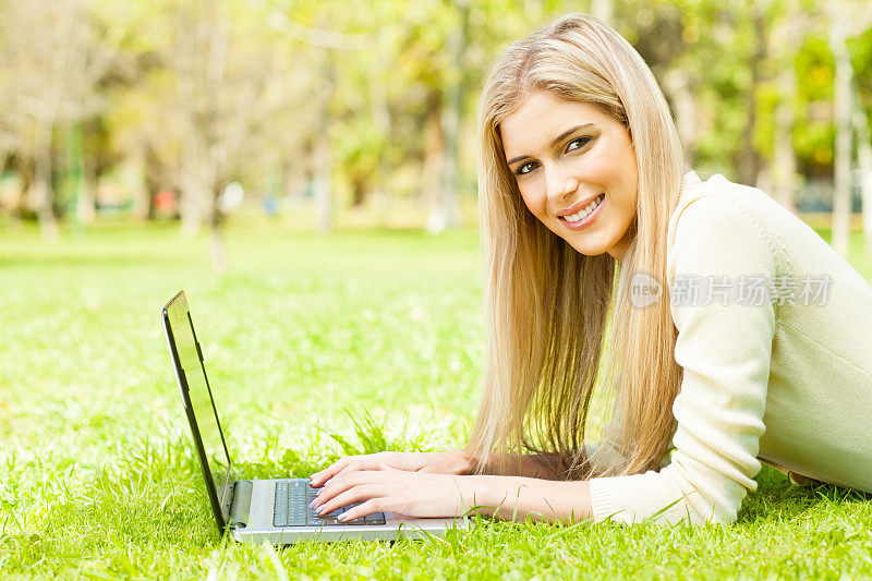 微笑的年轻女子在公园使用笔记本电脑。