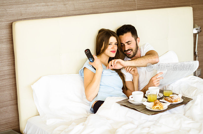 一对夫妇在床上吃早餐，看电视