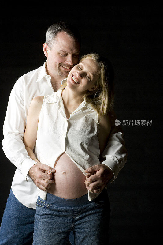 一对怀孕夫妇在黑色背景下拥抱在摄影棚里。