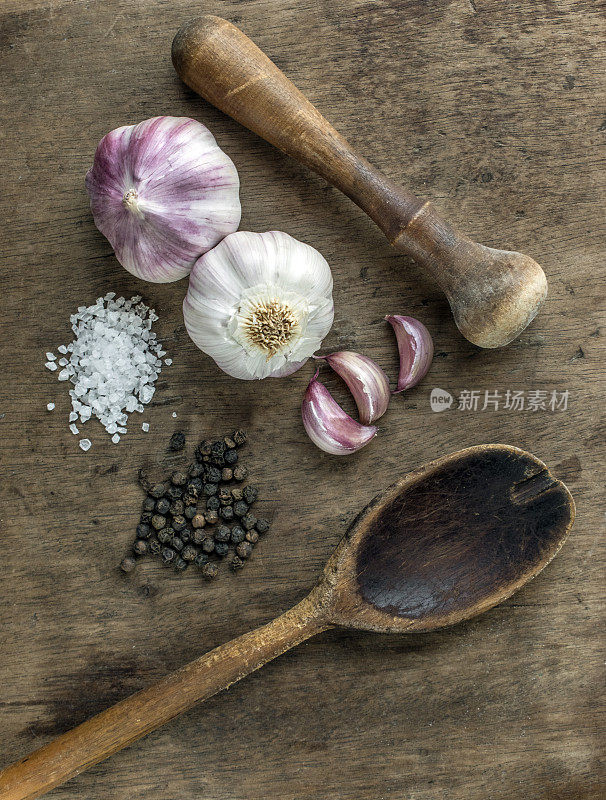 俯视图的香料和一个木制的烹饪勺子，研钵和杵，显示整个紫色的大蒜球，蒜瓣，黑胡椒和白色的盐，在一个木制的表面背景