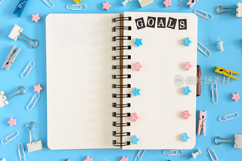 弹簧上的记事本是打开的。在食堂办公用品旁边。笔记本的白色页上写着“目标”，粉色和蓝色的星号。上面的照片，蓝色背景。