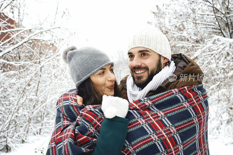 年轻的男孩在温暖的衣服拥抱和温暖她的女朋友在冬天的森林。年轻的恋人望着对方微笑