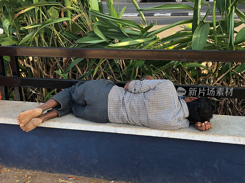 这是印度果阿邦帕纳吉海滩上的一名年轻印度教男子，他在午后的阳光下蜷缩着睡觉，在炎热的天气里午睡，打盹，疲惫不堪，赤脚在热带竹子和金属栅栏旁蜷缩着睡觉