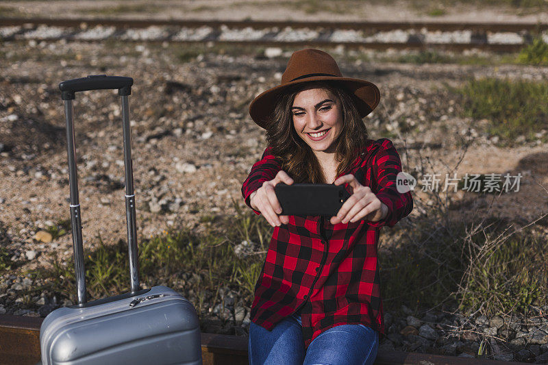 年轻漂亮的女子穿着休闲的衣服，带着行李箱和手机，面带微笑地走过铁路。户外生活方式。旅行的概念。她正在拍照或自拍