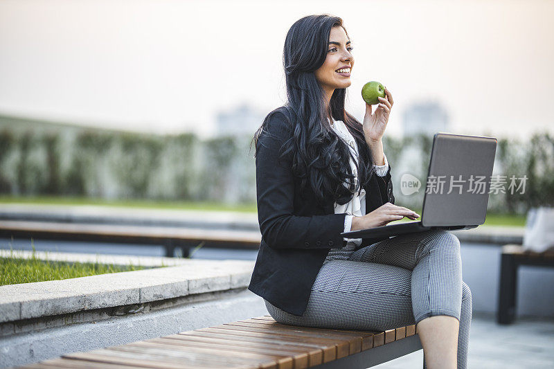女商人用手提电脑，在公园长凳上吃苹果