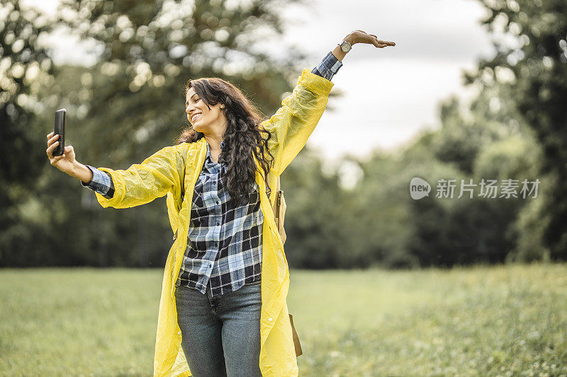 穿着黄色雨衣的年轻女子用智能手机自拍，微笑着享受和休息在自然中
