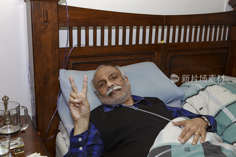 一名72岁的印度男子做完腹腔镜胆囊手术后躺在家中的床上恢复。他对着镜头做了胜利的V手势，表示他克服了困难。