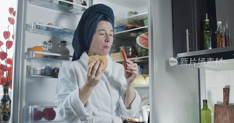 一个成熟的女人穿着浴袍和浴巾头巾在冰箱旁边吃着香肠和三明治