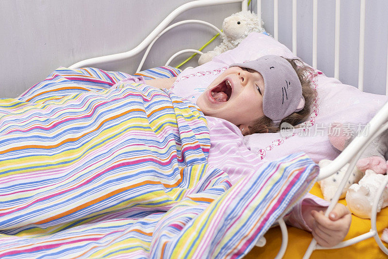 一个5岁的白人小孩，在婴儿床上伸懒腰、打哈欠。戴着面具睡觉的女孩。