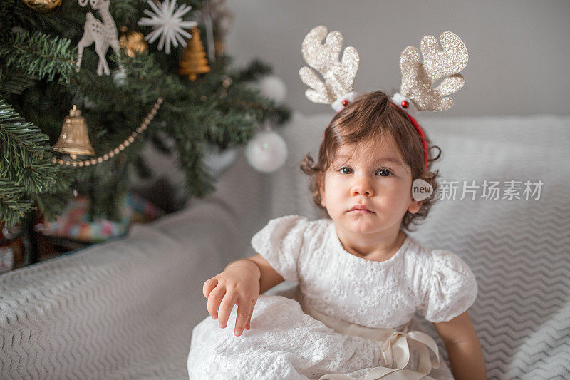 美丽的小女孩和鹿角坐在圣诞树下