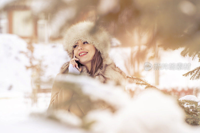 美丽的女人戴着皮草帽在冬天的公园里使用智能手机在下雪的天气下