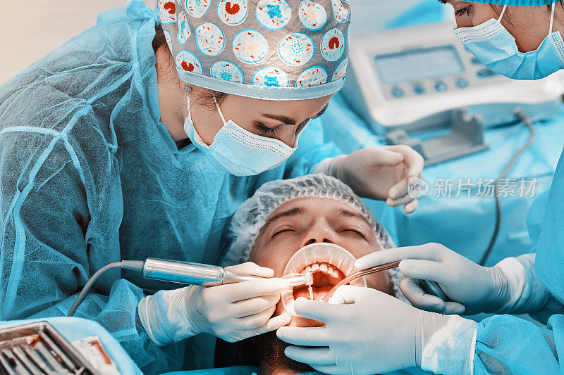 在一家牙科诊所，两名女牙医和牙科助理正在为一名穿着西装的病人进行治疗手术