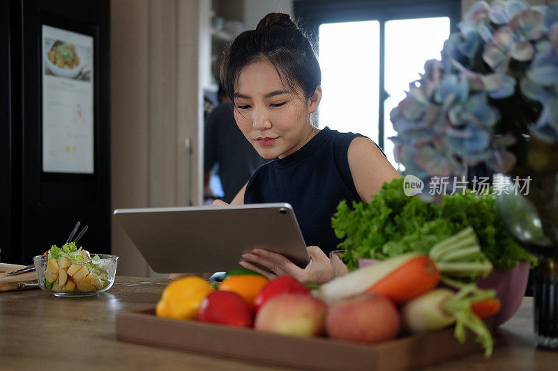 女人坐在厨房的餐桌上，餐桌上摆满了新鲜蔬菜，她在数字平板电脑上搜索菜谱。
