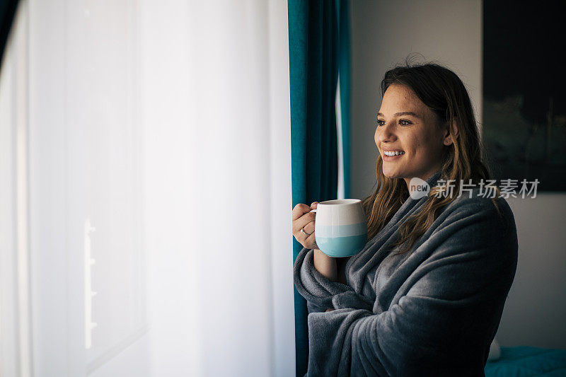 美丽的年轻女子裹着毯子站在窗户后喝茶