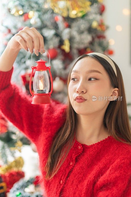 年轻美丽友好的亚洲女性女士穿着红色长袖毛衣衬衫，带着可爱的发带，拿着一个小户外灯笼烛光，装饰得很漂亮的圣诞树