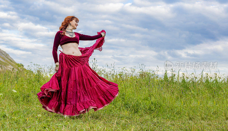 年轻的红头发女孩穿着红色的舞衣在碧绿的草地上跳舞，衬托着夏日的蓝天。