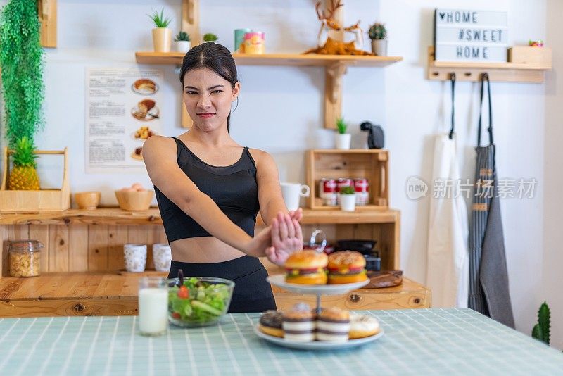 照片年轻美丽的亚洲女性女士在运动服装说不和拒绝诱惑汉堡和甜甜圈在厨房的桌子上，选择一碗沙拉与樱桃番茄和一杯牛奶的饮食和减肥健康的生活方式