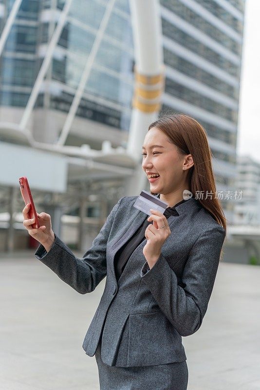 一名年轻的亚洲女商人穿着灰色的办公套装，在商业区用她的智能手机使用信用卡进行在线交易，她微笑着自信