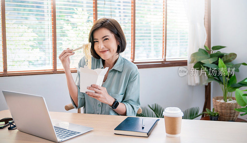 年轻的亚洲女性在办公室用笔记本电脑享受中餐的肖像，在家工作，女员工拿着筷子在工作场所吃外卖面盒饭。