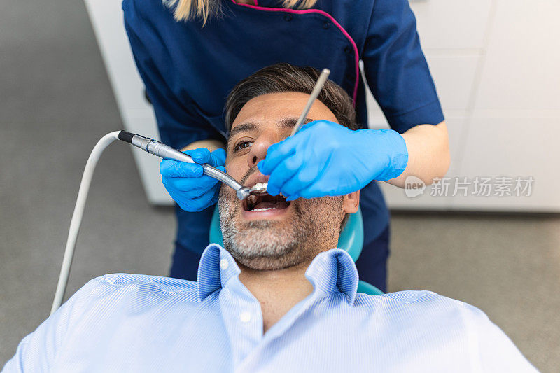 在现代牙科诊所，穿着蓝色制服的牙医正在给患者做牙齿手术，男性患者在治疗过程中
