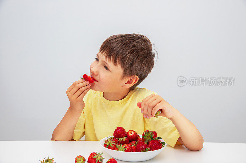 这个男孩吃成熟的红草莓。一个穿黄色t恤的孩子坐在桌子上，手里拿着草莓。美味健康的环保食品