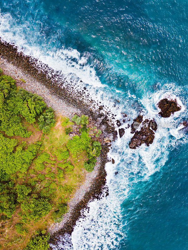 鸟瞰图:绿松石般的海浪撞击岩石海岸线，毗邻绿色海岸景观，郁郁葱葱，龙目岛。