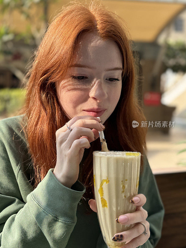 迷人的特写照片，红头发，年轻女子拿着灯罩杯香蕉奶昔，用纸吸管喝着不含酒精的饮料，户外咖啡馆餐厅花园，重点在前景
