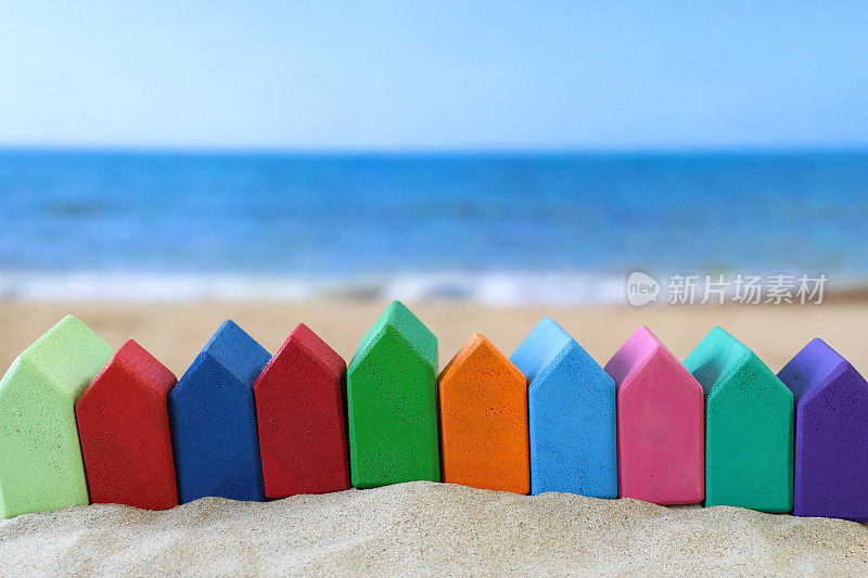 一堆沙子上的排彩虹木制模型度假小屋的特写图像，红色，橙色，浅蓝和深蓝，粉色，浅绿和深绿，紫色，沙滩背景，退潮时的波浪，海岸线，度假租赁和度假屋的概念