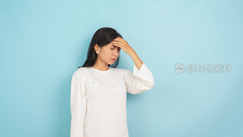白衣亚洲女性，闭着眼睛，带着痛苦的表情，按压太阳穴，在浅蓝色背景下显示出头痛或偏头痛的迹象