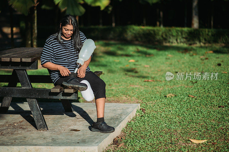 在阳光明媚的日子里，一名亚裔印度残疾人微笑着坐在公园长椅上调整她的义肢