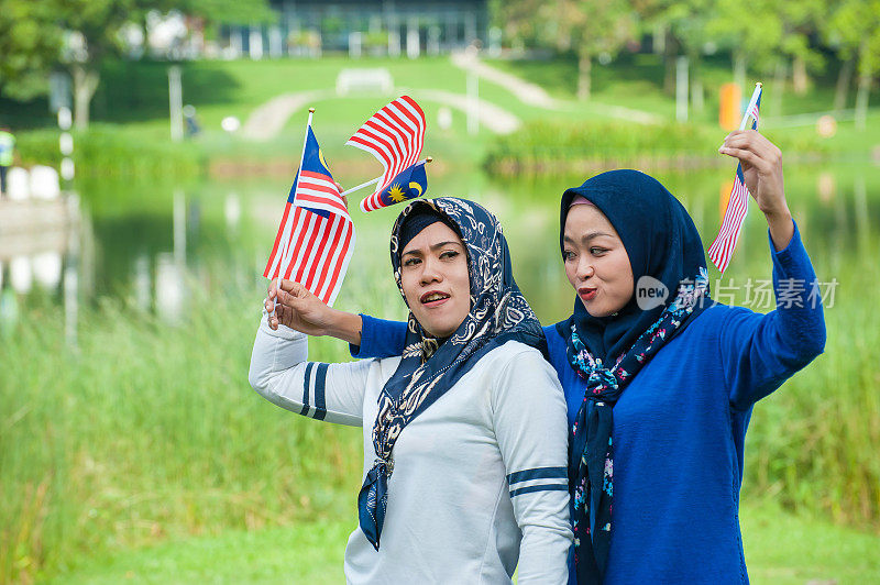 两名年轻的马来西亚妇女戴着头巾，挥舞着马来西亚国旗，以高度的爱国主义精神在公园庆祝独立日