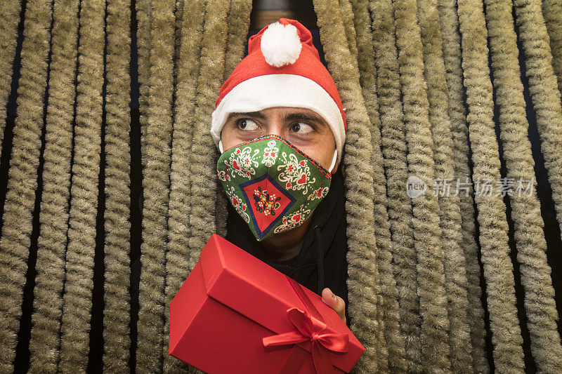 年轻男子的肖像戴着防护面具和圣诞帽和拿着圣诞礼物通过前门