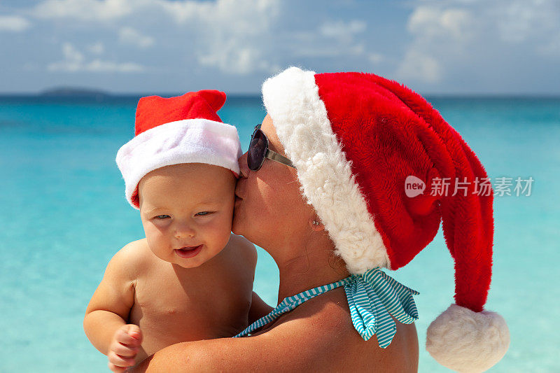 妈妈抱着快乐的婴儿戴着圣诞帽在加勒比热带海滩上