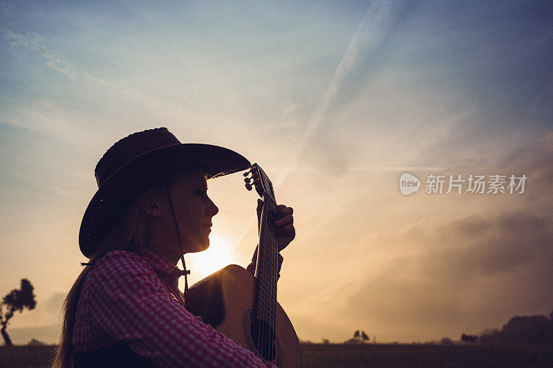 一个浪漫的女牛仔在户外玩吉他的低调肖像在日出