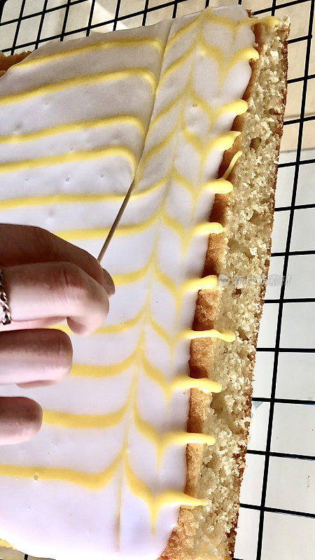 图片:一个不认识的人用鸡尾酒棒在白色方糖糖霜上画出黄色柠檬味糖霜的羽毛图案，自制的柠檬托盘烤蛋糕放在金属冷却架上，高架视野