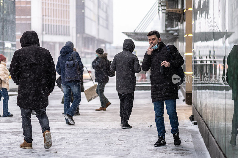下雪时，一名戴着医用口罩的男子在街上与路人一起吸烟