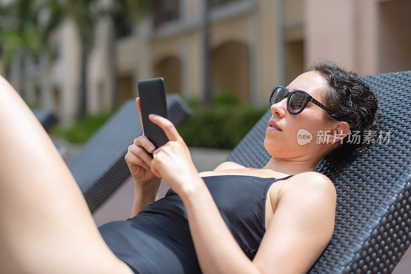 年轻的成年女性在豪华度假村日光浴时使用平板电脑