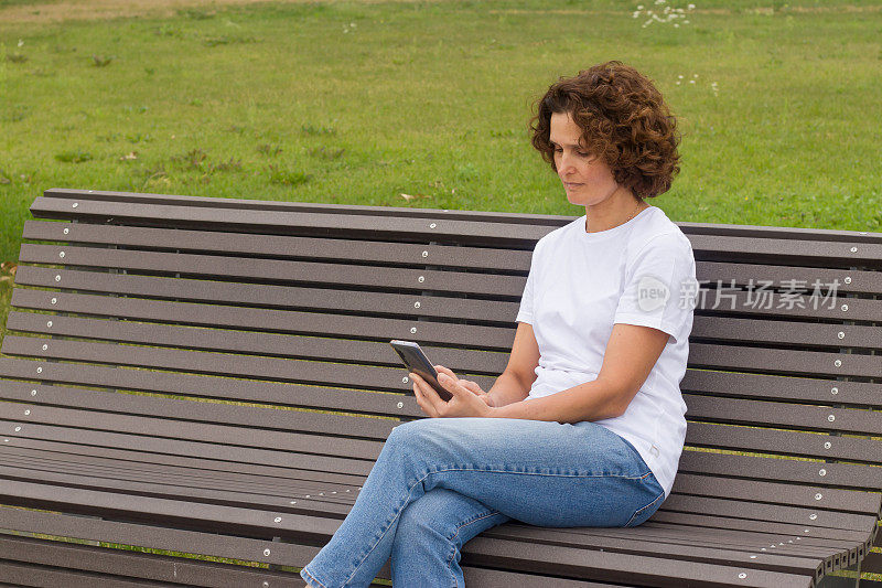一位45-50岁的妇女坐在长凳上，手里拿着一部手机