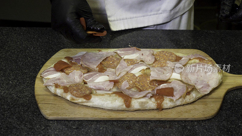 穿着黑色烹饪手套的厨师躺在木板上制作披萨，美味的食物概念。框架。手把橄榄和肉放在披萨上的特写。
