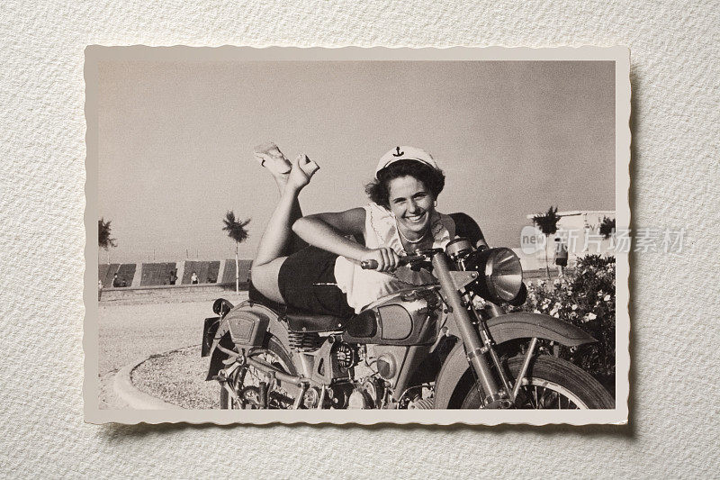 微笑的女孩骑着摩托车在海边。50年代的照片。