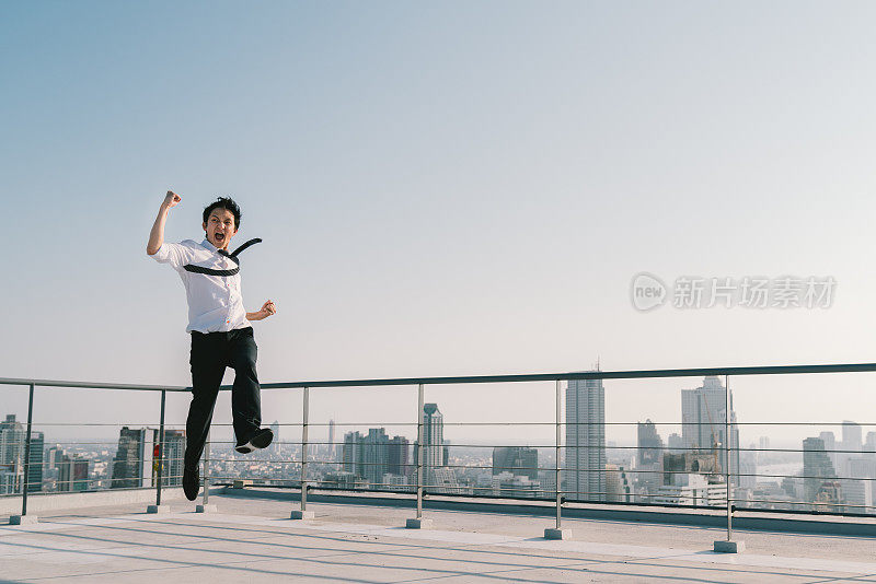 年轻英俊的亚洲商人跳高，庆祝成功获胜的姿态在建筑屋顶。工作、工作或成功的商业理念。城市景观背景与拷贝空间在阳光明媚的蓝天