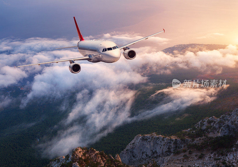 美丽的飞机在日落时飞过低空的云层。景观有客机，岩石，森林，蓝色的天空和黄昏的橙色阳光。客机。商务旅行。商用飞机