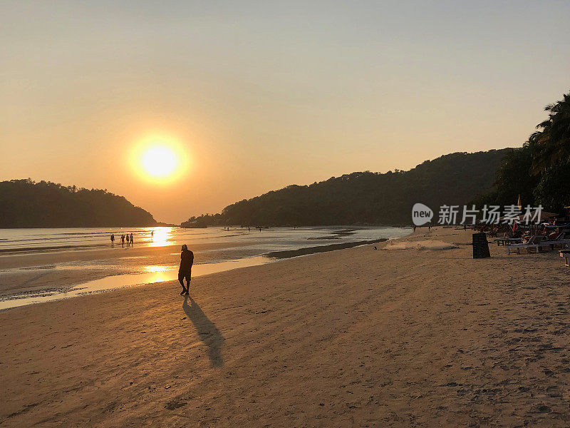 图片中，一名穿着短裤的男子走在印度果阿的帕洛伦海滩的海岸线上，夕阳下，海浪拍打着海滩