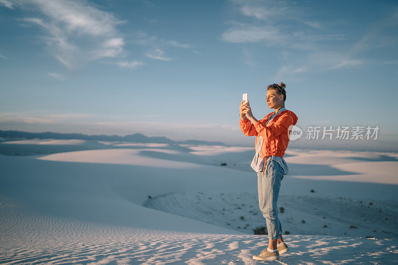 年轻女子在白沙沙漠用智能手机拍摄以风景为背景的自拍，时髦女孩站在沙丘上欣赏美国国家纪念碑的美丽风景