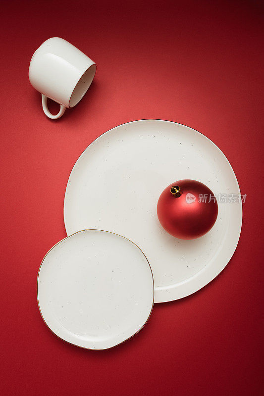 有创意的餐桌布置，有两个盘子，一个杯子，红色背景上有鲜红色的圣诞装饰物。最小限度的庆祝想法。